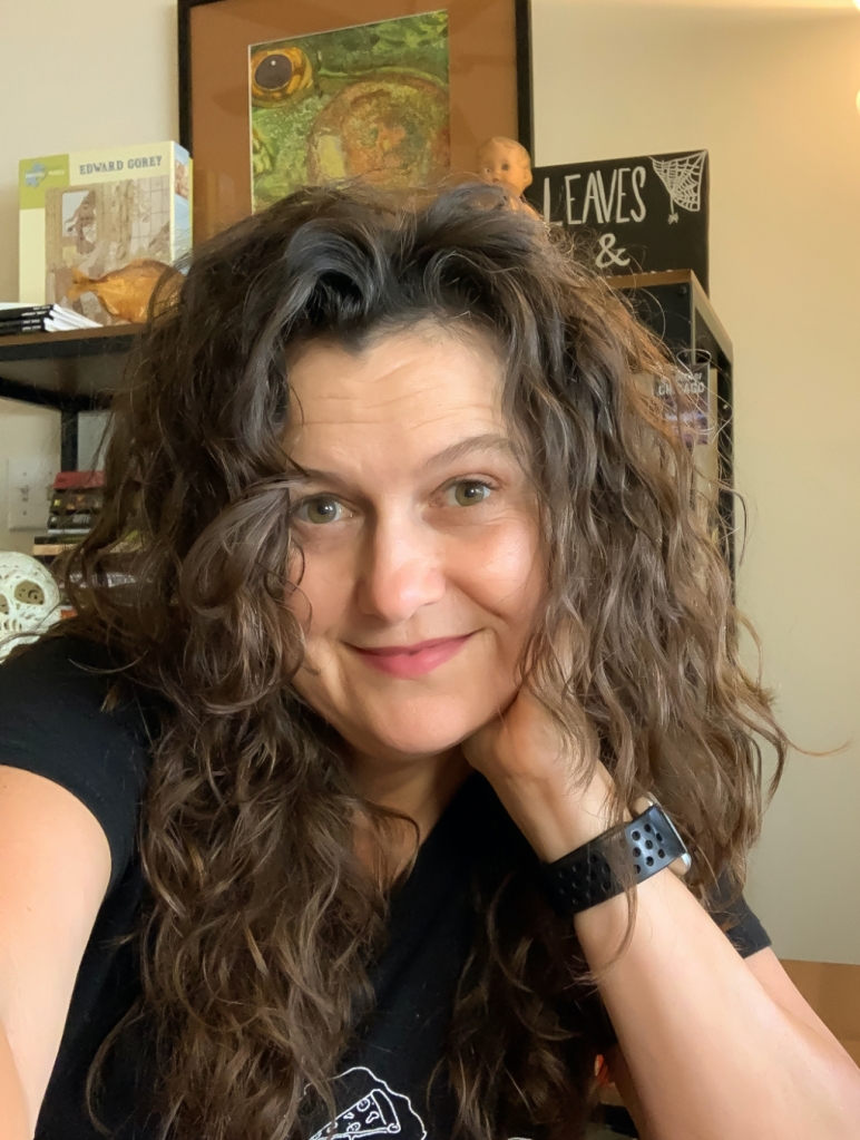 Photo of Victoria Nations, a woman with hazel eyes and curly brown hair. She is smiling and leaning on hear hand. A bookshelf with a skull, babydoll, stuffed piranha, Edward Gorey puzzle, and horror fiction is behind her.